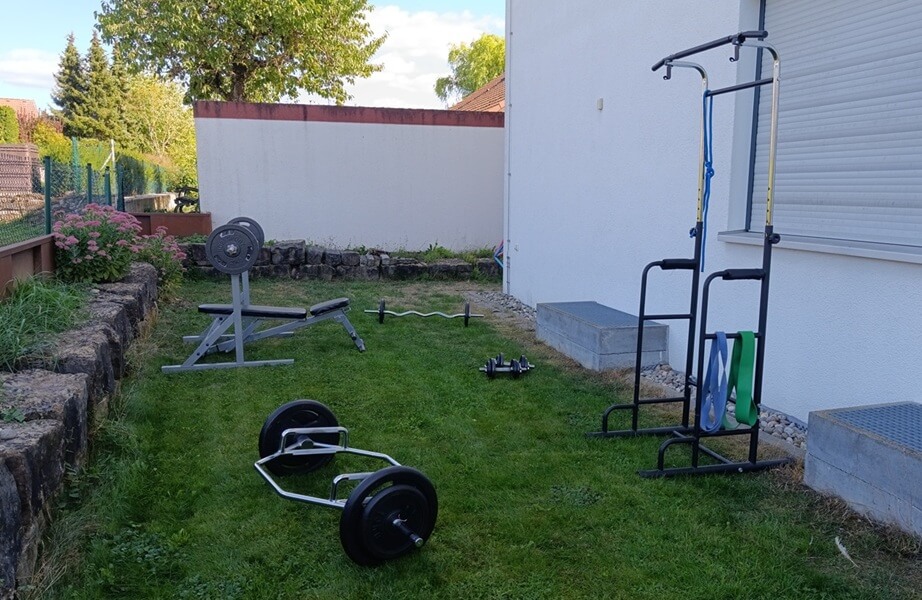 Der VaterFit Trainingsgarten für mein Personal Training Angebot in Bad Friedrichshall. Zu sehen sind ein Klimmzugturm mit Widerstandsbändern, eine Hexbar, eine Hantelbank, eine Langhantel in einer Ablage, eine SZ-Stange sowie zwei Kurzhanteln.
